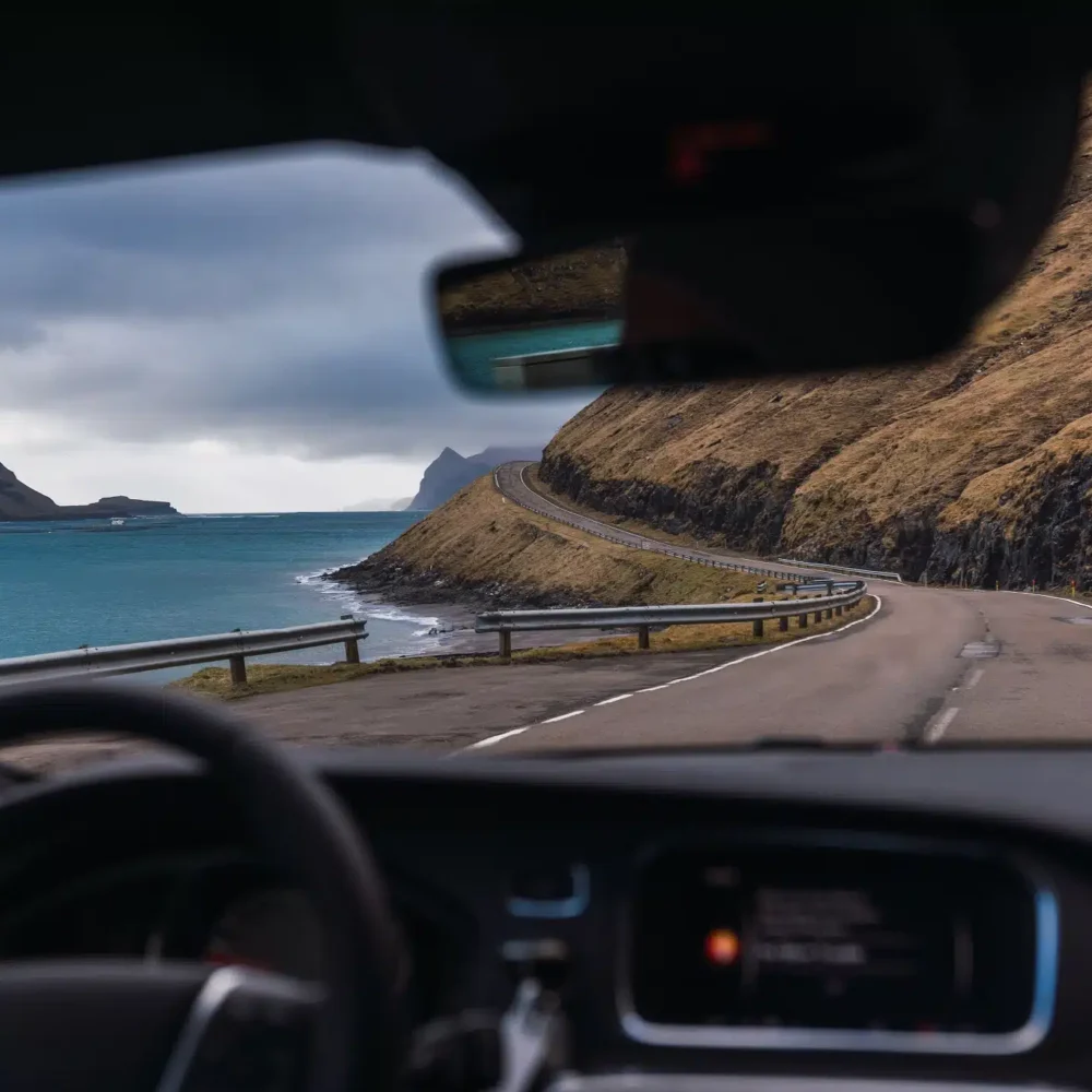 Driving a rental car along a scenic coastal road in the Faroe Islands with mountains and ocean views