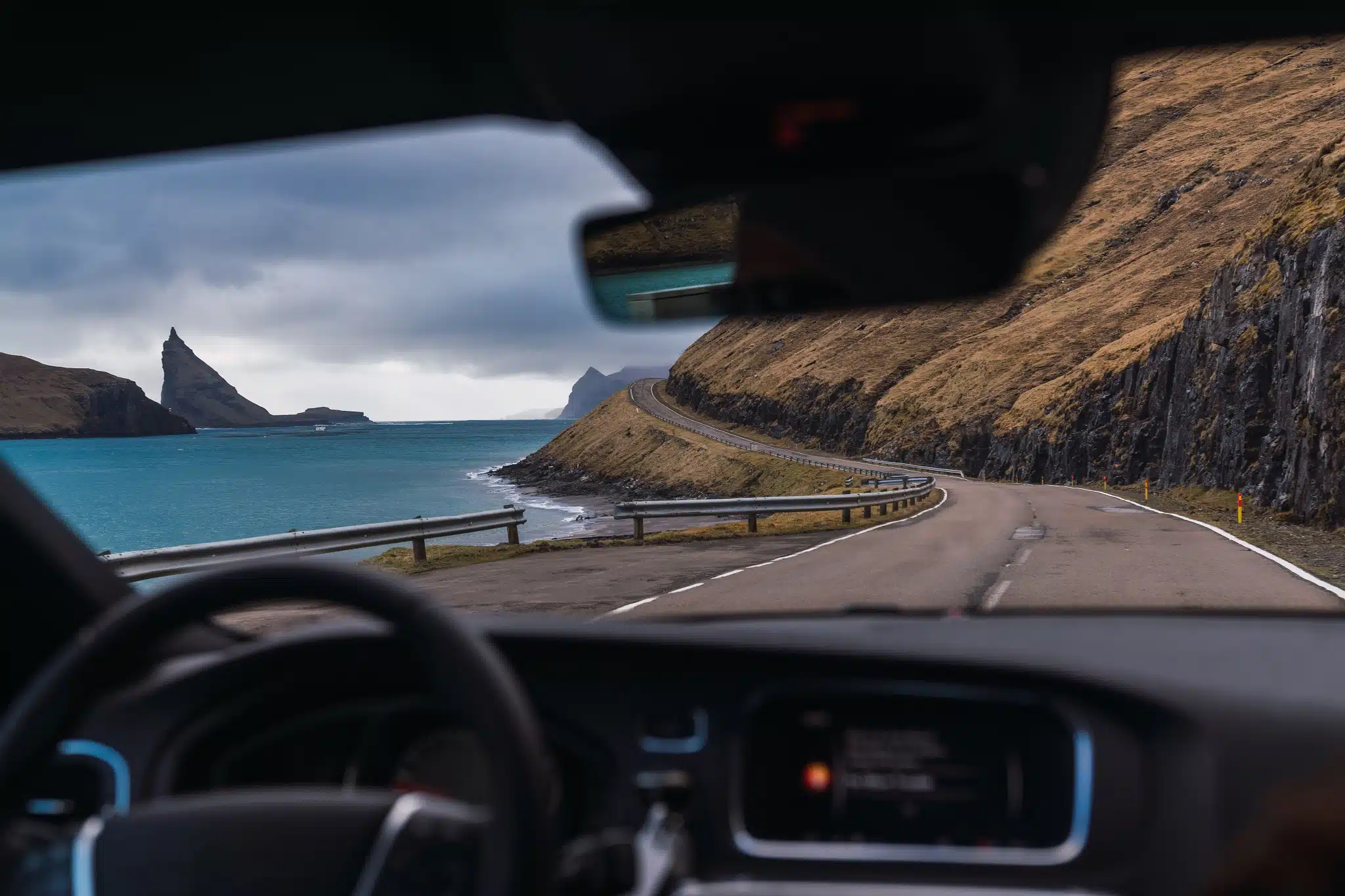 Driving a rental car along a scenic coastal road in the Faroe Islands with mountains and ocean views